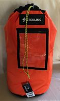 Front view showing orange color, clear ID window, brand label and drawstring closure with tag attached.