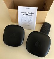 Photo of two black wireless doorbell receivers, one black push button transmitter, user manual and cardboard box.