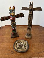 Front view of two totem poles (one painted, one carved natural finish) and a small pottery dish with carved symbols placed on a wooden surface.