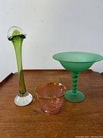 Front view of three art glass pieces on wood surface against white background: tall green chanterelle vase, green frosted compote with hobnail stem, small pink swirl bowl