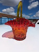Ruby to amber colored art glass brides basket with ruffled top edge and solid yellow handle outdoors on a white surface.
