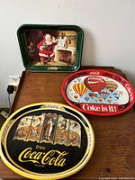 Photo showing all three Coca-Cola trays arranged on a table: green Santa tray, red balloon tray, and black oval tray with vintage women images.