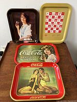 Set of 4 vintage enameled metal Coca-Cola advertising serving trays, showing varied images and colors, laid together on table.