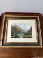 Frontal view of the wood framed alpine landscape painting showing the scene with mountains, river, and valley, along with the artist signature.