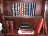 Two wooden shelves inside a glass-front book cabinet filled with hardcover books in black, red, and blue bindings. Bottom shelf shows a stack of several books.