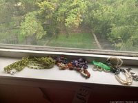 Photo of multiple necklaces displayed on a window sill, showing variety of stones and beads including green beads, pearls, and multi-colored semi-precious stones.