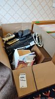Overhead photo of open box containing VCR, cordless phones, RCA radio in wooden cabinet, coffee cup warmer, and other items.