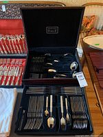 Open black presentation case showing stainless steel cutlery including forks, knives, spoons, and serving utensils organized in velvet compartments with Wexford Forge brand on inside lid.
