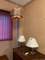 Photo showing all three lamps: two table lamps on a wooden surface, and one hanging lamp with overlapping wooden panels suspended from ceiling chain.