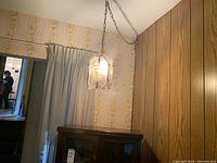 Hanging plug-in light fixture with glass panels suspended from a chain in a room with floral wallpaper and a wooden cabinet below.