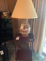 Front angled view of the MCM table lamp on a wooden two-tier side table, showing ceramic base with mottled finish and pleated cream cloth lampshade.
