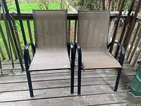 Photo of two metal frame outdoor chairs with beige/brown mesh seats and backs positioned on a wooden deck near railing and greenery.