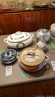 Photo showing lot items on table including lidded serving dishes, Delft bottle, and Milne pot