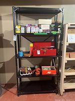 Front view of black metal shelving unit with various items on shelves such as cardboard boxes, a red toolbox, and an orange extension cord.