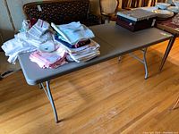 Photo shows whole tabletop with folded linens and decorative boxes on top, the table top is gray plastic with faint texture and rounded corners, supported by four metal legs.