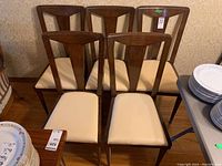 Five vintage wood dining side chairs arranged in two rows showing cream padded seats and trapezoidal backrests with dark wood frames.