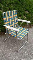 Side perspective showing colorful woven plastic strap seat and backrest with aluminum frame and white plastic armrests.