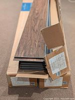 Two boxes of AduraMax vinyl plank flooring in Sundance pattern, Gunstock color, with planks laid on top showing wood grain detail.