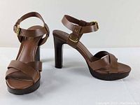 Front and side view of pair of brown Lauren Ralph Lauren women’s heeled sandals with ankle straps and gold buckles on a white background.