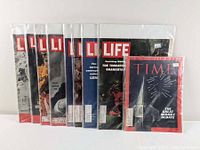 Nine magazines including eight Life magazines and one Time magazine in plastic sleeves arranged standing vertically on a flat surface
