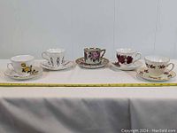 Full view of all five teacups and saucers arranged in a line on a white surface showing the variety of floral patterns and gold trim.
