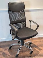 Front-angle view of the black office chair showing mesh backrest, padded seat, plastic armrests, and chrome base with rubber casters on hardwood floor.