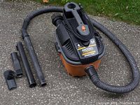 Rigid Professional 4 gallon shop vacuum with orange base and black top, hose connected, and multiple attachments laid out beside it on pavement.