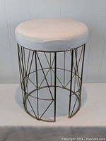 Front oblique view of round cream fabric cushioned vanity stool with geometric gold tone metal base on white surface against lined wall.