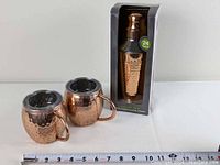 Shaker in box and two copper plated mugs with hammered texture and stainless steel interior, on flat white surface next to measuring tape for size reference
