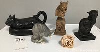 Five cat collectable items displayed together including a black cat butter dish, a gray cat figurine, a carved curled cat figure, the Fremiet Cat sculpture and the Joe Slockbower Mountain Lions sculpture.