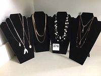 Eight gold-tone necklaces displayed on black velvet necklace busts in two rows. Variety of chain types and pendant styles including beads, feathers, and medallions.