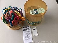 Photo showing 20 colorful Kraft edge scissors placed in a round hardwood rotating rack next to a pattern guide and a plastic container.