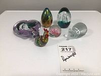 Full view of all six paperweights on a white surface including heart, egg shapes, mouse, rooster, and floral design with auction tag in front.