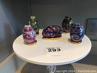 Five Laurel Burch cat collector items displayed on a white round table. Four upright cat figurines in various colors and one large cat-shaped egg in front. Each item sits on a gold-tone base.