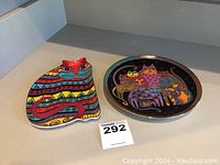 Two cat-themed decorative plates displayed side by side on a shelf. Left is a uniquely-shaped Laurel Burch colorful cat plate, right is a round Franklin Mint plate featuring multiple cats.