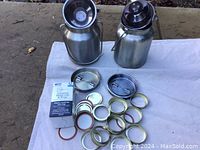 Two stainless steel Lindy's milk pails with lids and various lid parts laid out on a white cloth against an outdoor background, showing full items and all components.