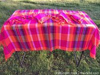Image shows bright pink, orange, and red striped hand-woven tablecloth displayed on a metal frame outdoors.