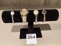 Display of three ladies' watches on a black stand, including a visible detailed gold-tone skeleton watch band, two smaller silver-tone watches with clasp bands.