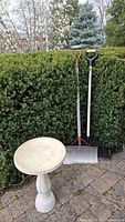 Outdoor scene showing white plastic bird bath alongside two shovels propped against a hedge on a stone patio.