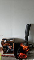 Photo of Black & Decker BV5600 blower with original box and HS1012 trimmer, both orange and black, placed on the floor against a wall.
