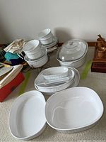 Overview of all items in lot including white Corning Ware casserole dishes with lids, serving stands, and spoon rest on carpet floor.