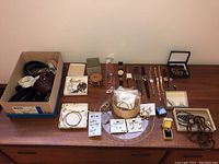 Lot overview showing belts in box on left, costume jewelry including earrings, bracelets, necklaces, and watches arranged on table