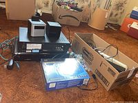 Lot contents with various electronics including alarm clocks, players, router, and power bar shown on floor and in box.