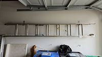 Photo showing a metal extension ladder mounted on a garage wall above trash bins and storage items.