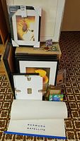 View of various frames in different sizes stacked against wall with protective corner covers, including flower-themed prints and large rolled poster labeled 'BERMUDA SATELLITE'. Small boxed pins on top of boxes.