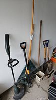 View of the garden tools showing rakes, shovels, line trimmer, and garden hoe standing on the floor against a wall in a garage.