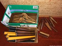 Box containing wooden table legs in various sizes and colors, all with screw fittings for use as furniture legs.