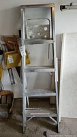 Front view of silver aluminum step ladder with four steps and support bracket in garage setting.