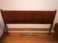 Full view of teak wood headboard with metal frame positioned against a white wall on hardwood flooring.