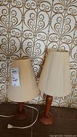 Two mid-century modern teak lamps with pleated cream shades, one slightly taller. One lamp has a bulb, the other does not. Electrical cords visible. Both lamps approximately 19 inches tall.